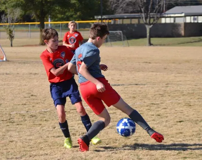 Two soccer players in red and blue contest the ball on a grassy field.