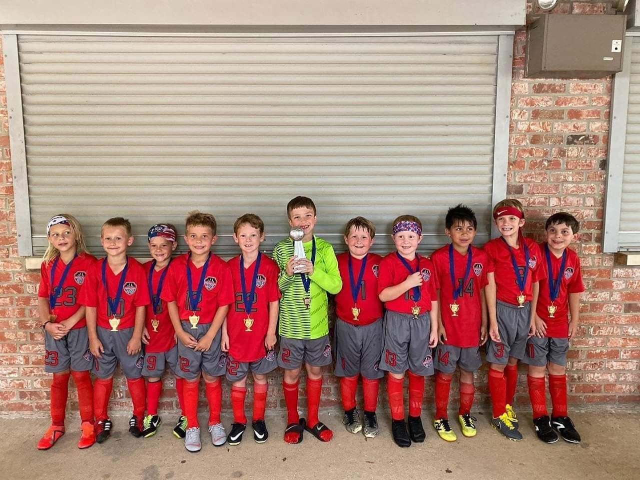 Group of children in red soccer uniforms standing in front of a closed garage door.