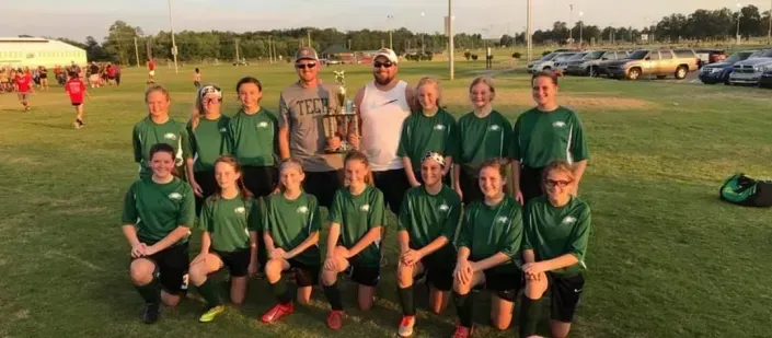 Youth soccer team posing on a field at sunset, wearing green jerseys with coaches behind them.