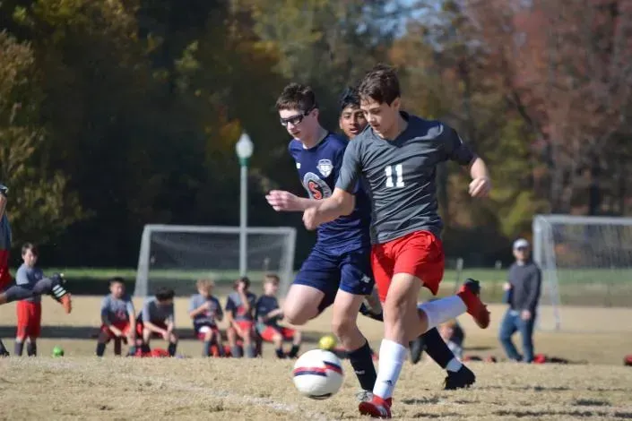 Two soccer players in blue and gray jerseys chase the ball on a grassy field with goalposts in the background