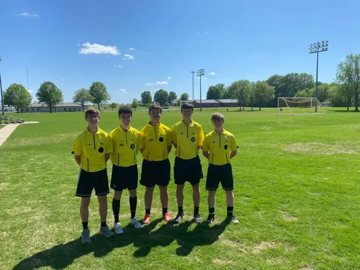 Five referees in yellow shirts stand on a grassy soccer field in bright sunlight.
