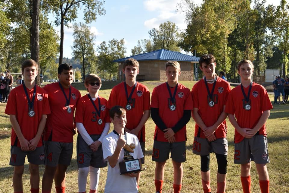 Youth sports team in red jerseys posing outdoors, with a child holding a trophy in front