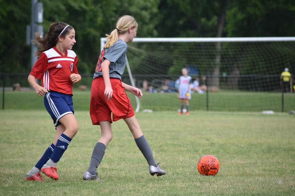Two soccer players chase an orange ball on a grassy field with a goal in the background