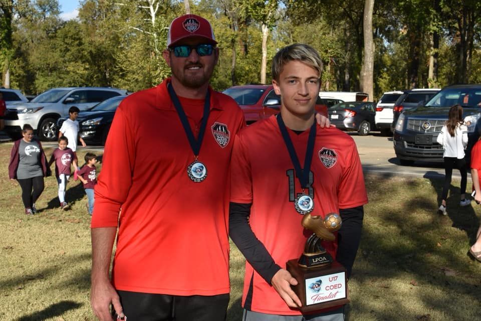 Two men in red shirts and medals pose outdoors, one holding a trophy.