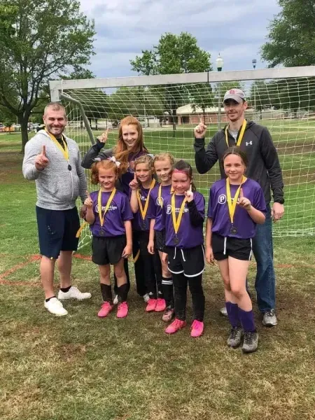 Group of children and adults posing outdoors on a grassy field, smiling with medals and a trophy