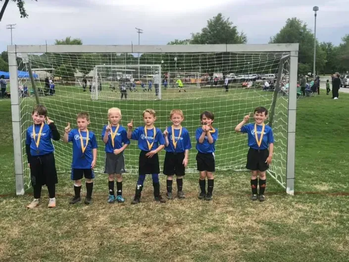 Youth soccer team posing in front of a goal, wearing blue jerseys and medals on a grassy field.