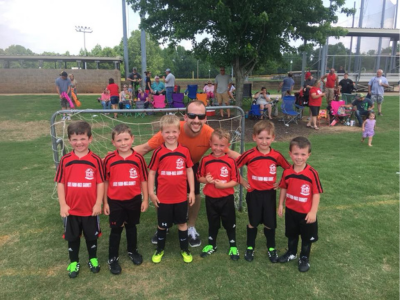 Youth soccer team in red jerseys posing with a coach on a grassy field, with goals and spectators behind them.