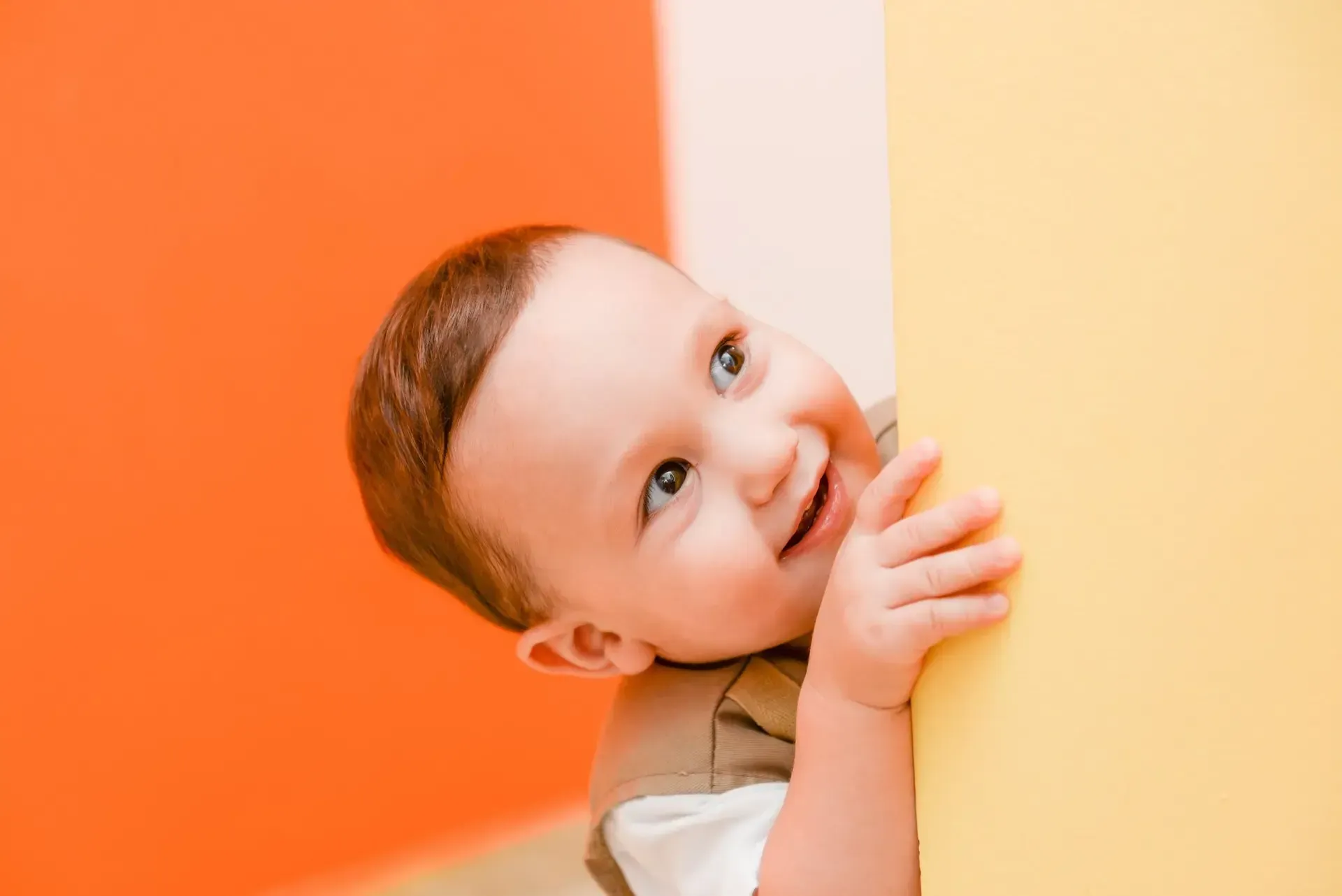 Smiling baby in family assessment centre peeks around a yellow wall, orange background.