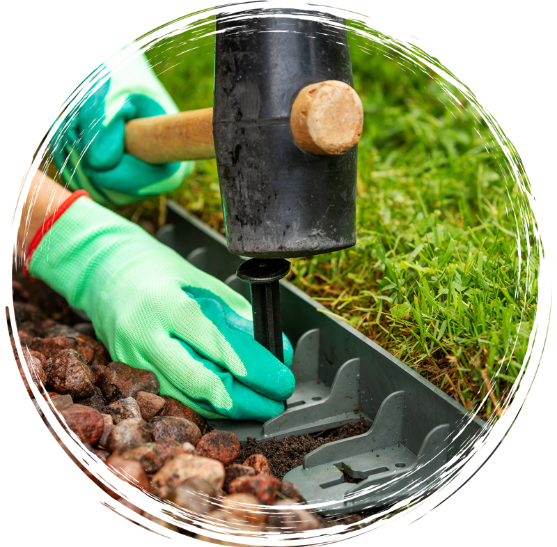 Green-gloved hand using a mallet to drive a garden edging stake into soil beside grass and rocks
