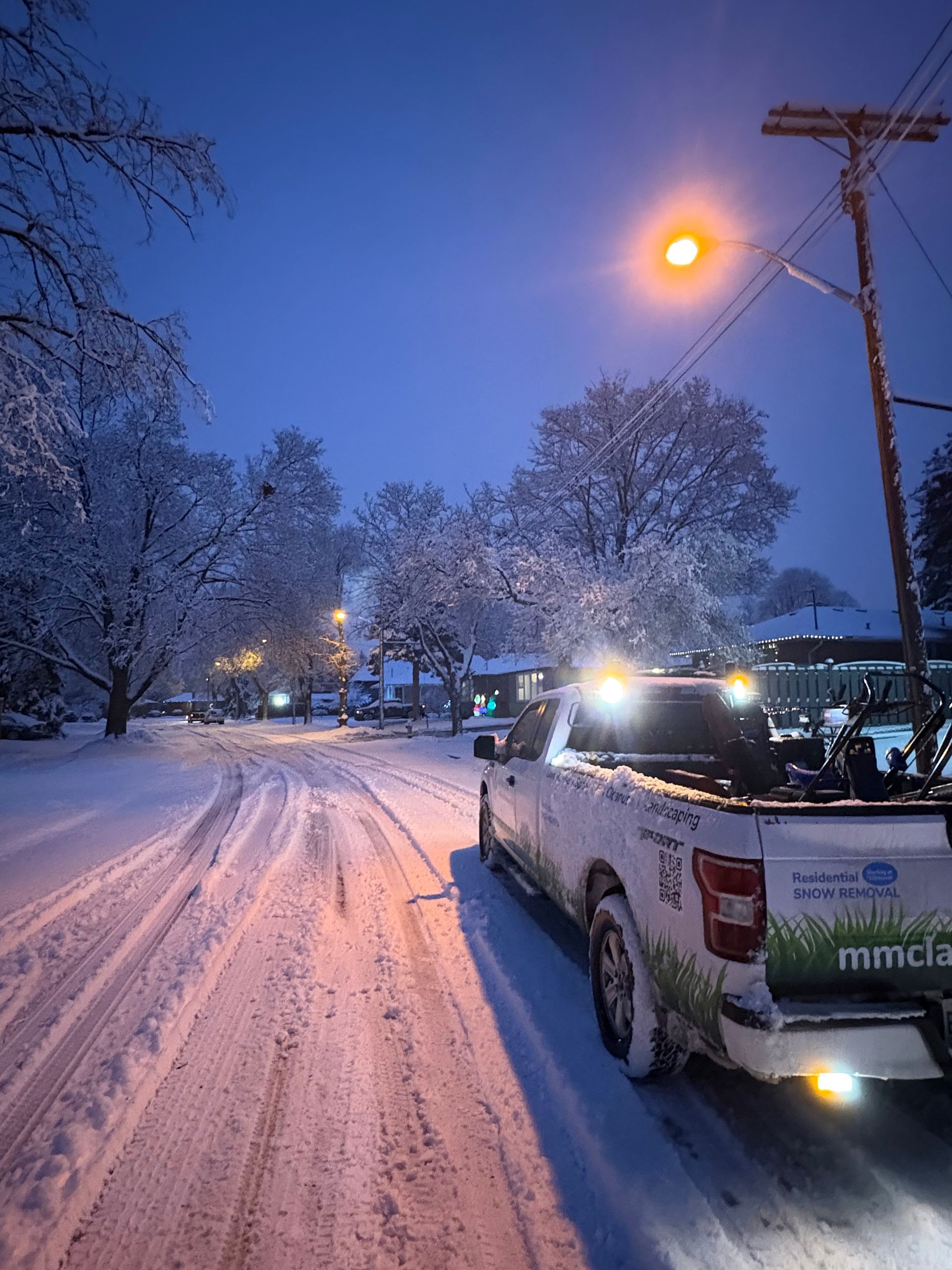 Snowy street at dusk with a white utility truck parked under glowing streetlights and snow-covered trees.
