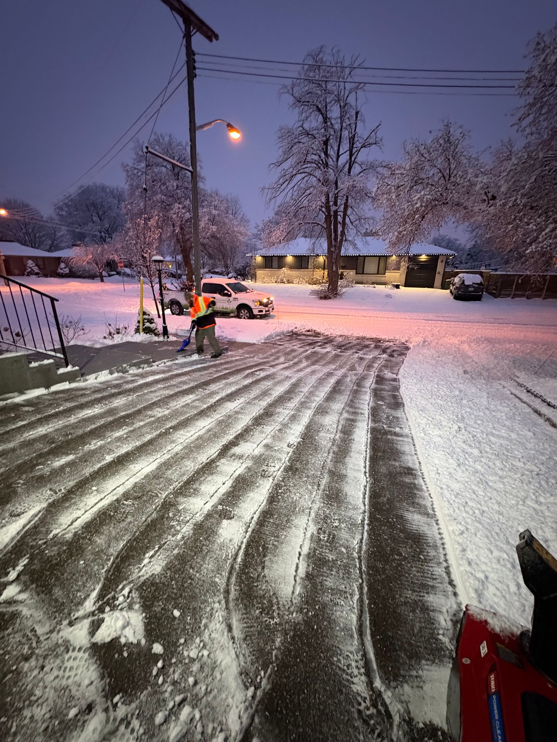 Snow-covered driveway at dusk with a snowblower clearing a path in a residential neighborhood.