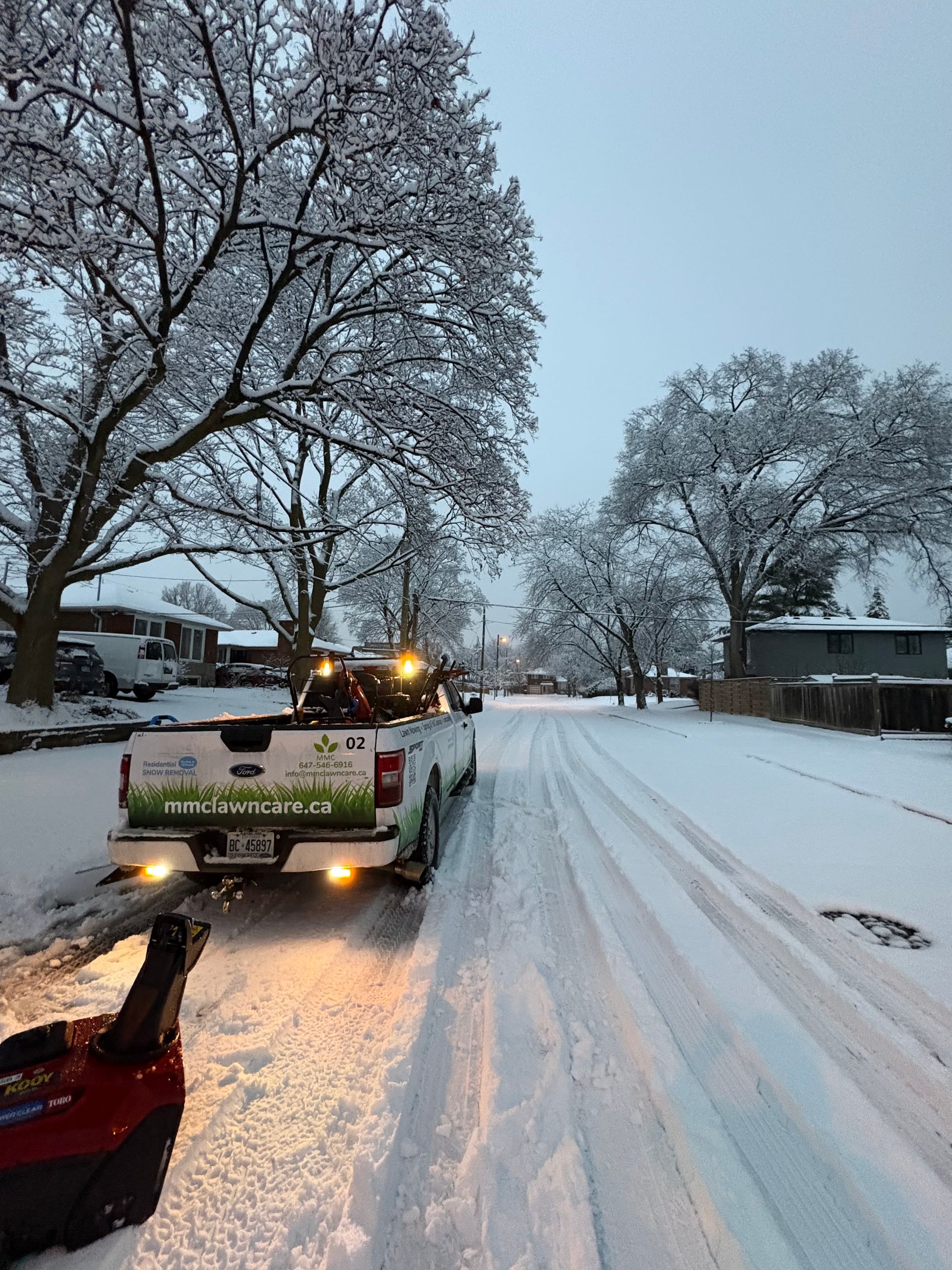 Snow-covered residential street with a pickup truck plowing snow under a gray winter sky