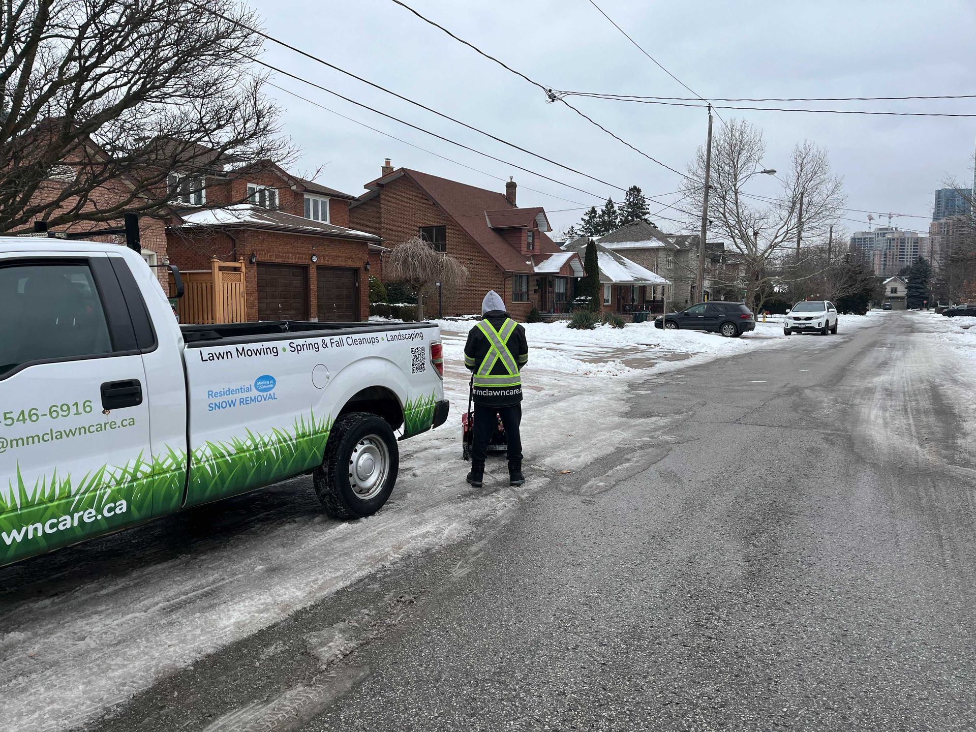 Person in a reflective vest using a snowblower on a snowy street, white truck with green logo nearby, houses in background.