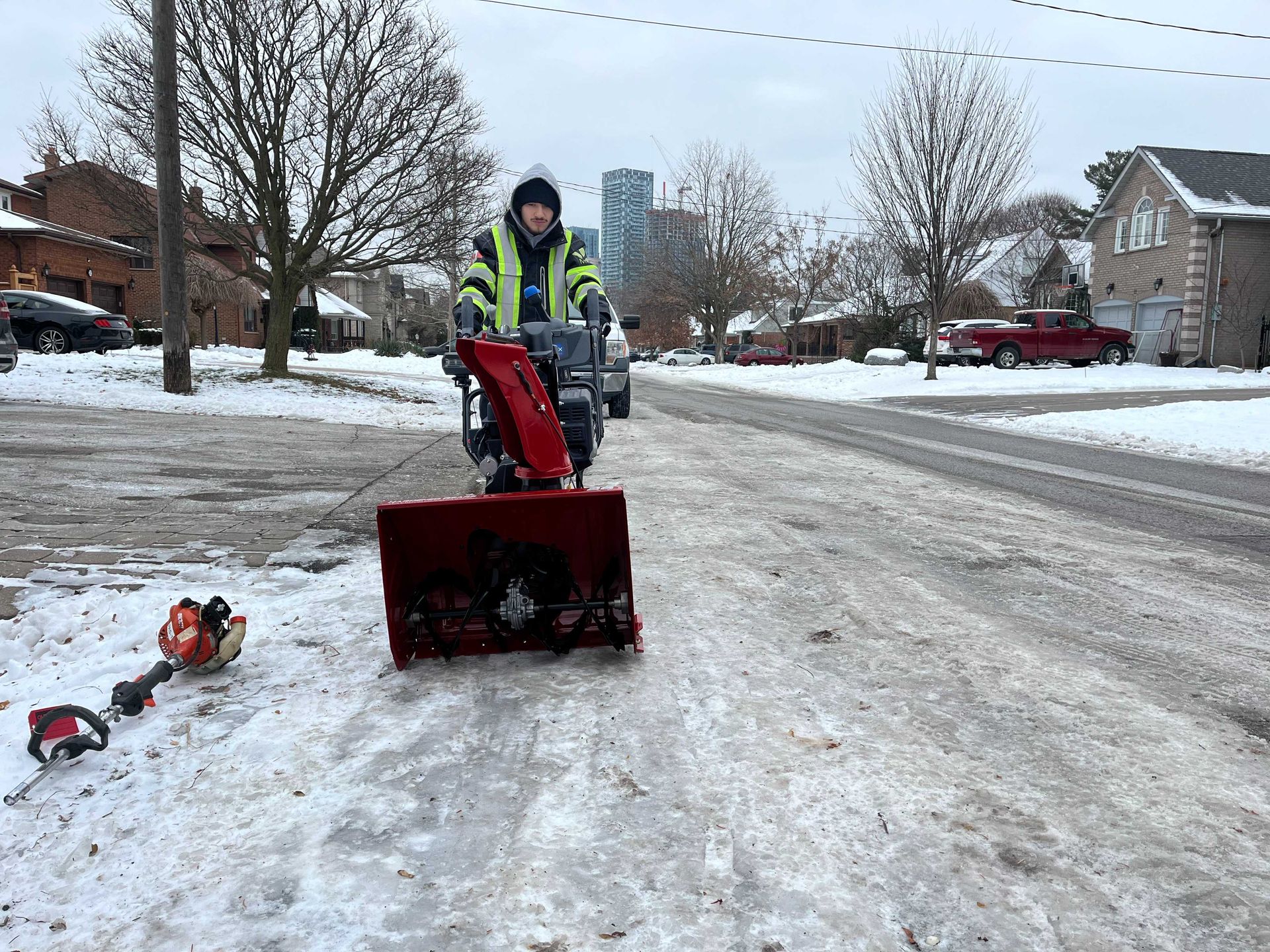 Person using a snowblower on a snow-covered street in a residential area. A string trimmer lies in the snow.
