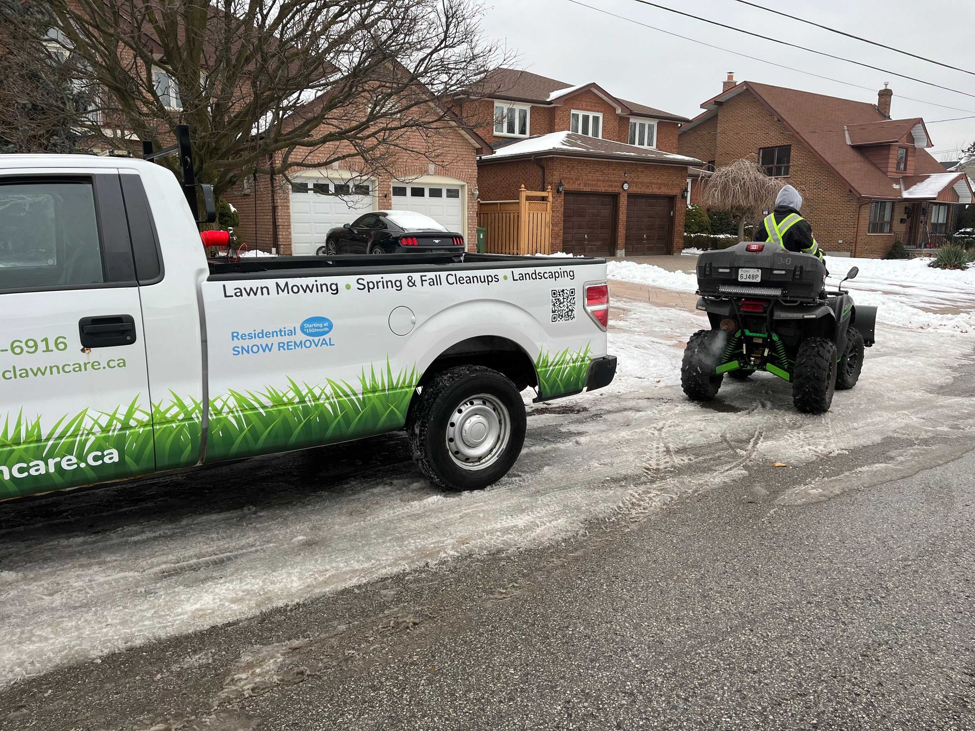 White pickup truck with green grass graphic towing an ATV on a snowy street in front of houses.
