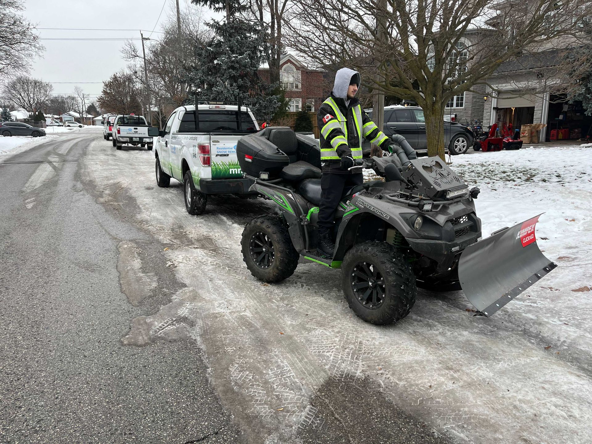 Person on ATV with snowplow clearing a snowy street. A truck with a trailer is behind the ATV.