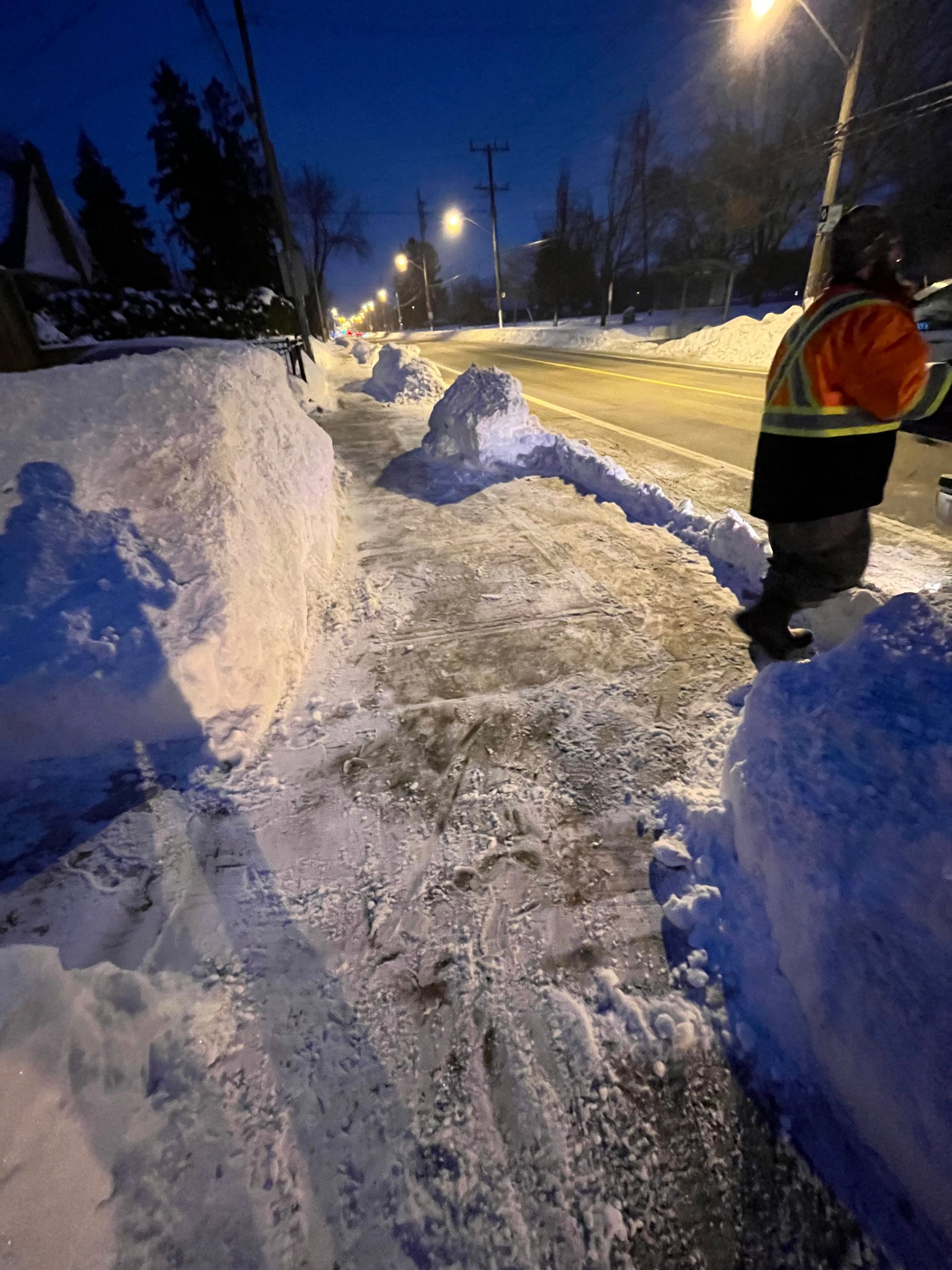 Snow-covered street at dusk with tall snowbanks and a worker in an orange jacket clearing the path