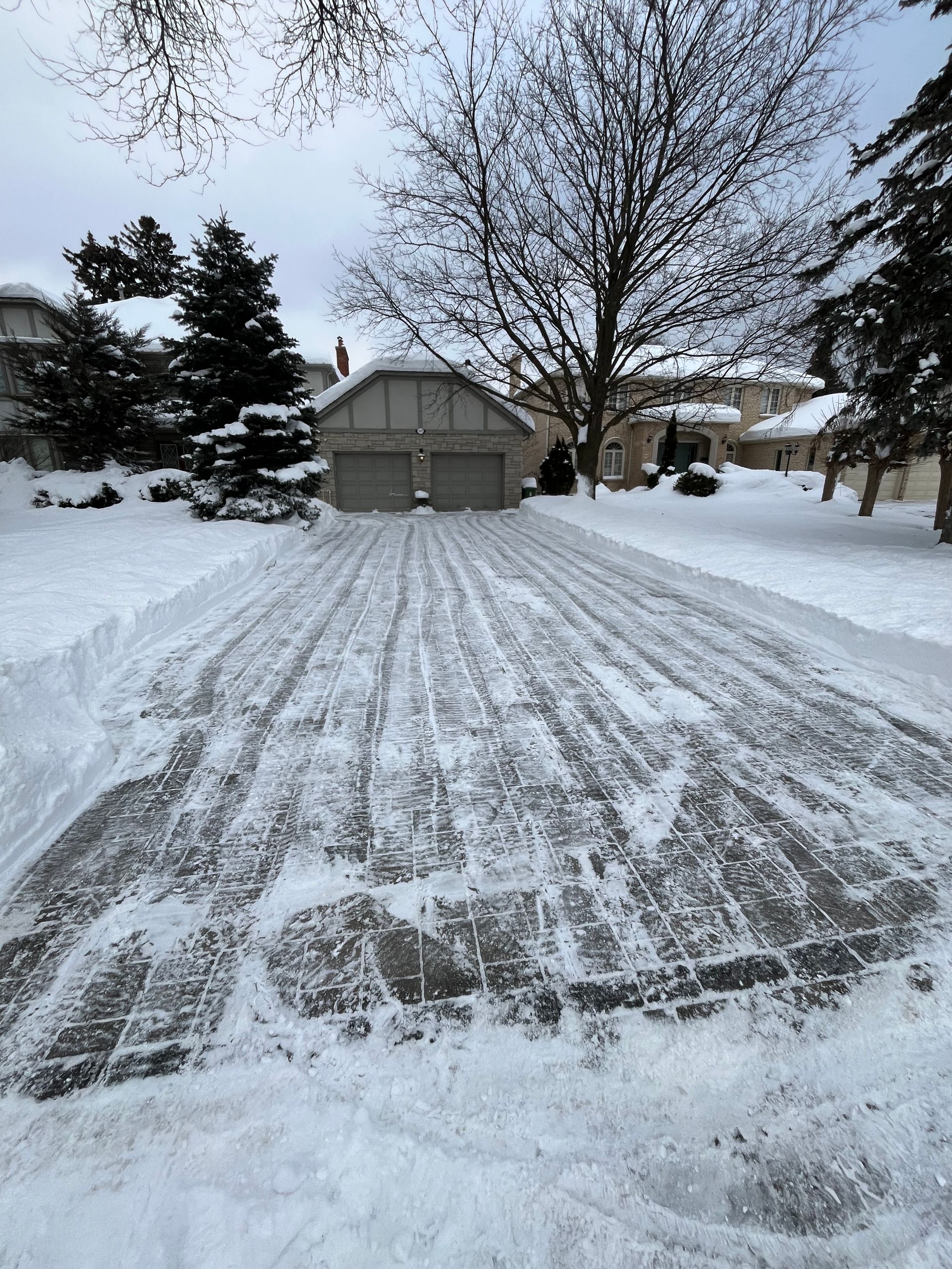 Snow-covered driveway leading to a gray garage in a suburban yard with bare trees.