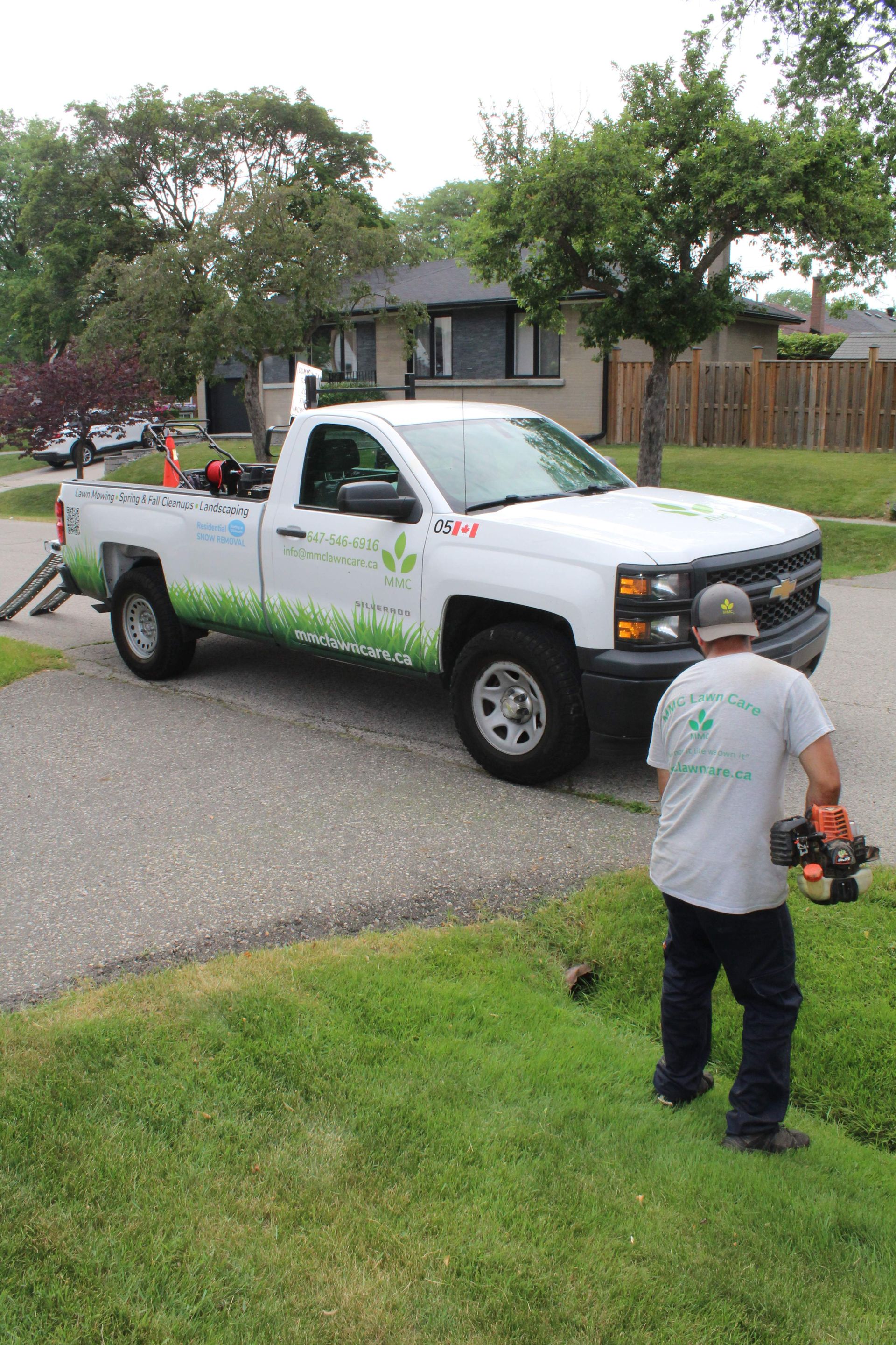 White pickup truck parked by a suburban street, with a person using a leaf blower on the lawn.