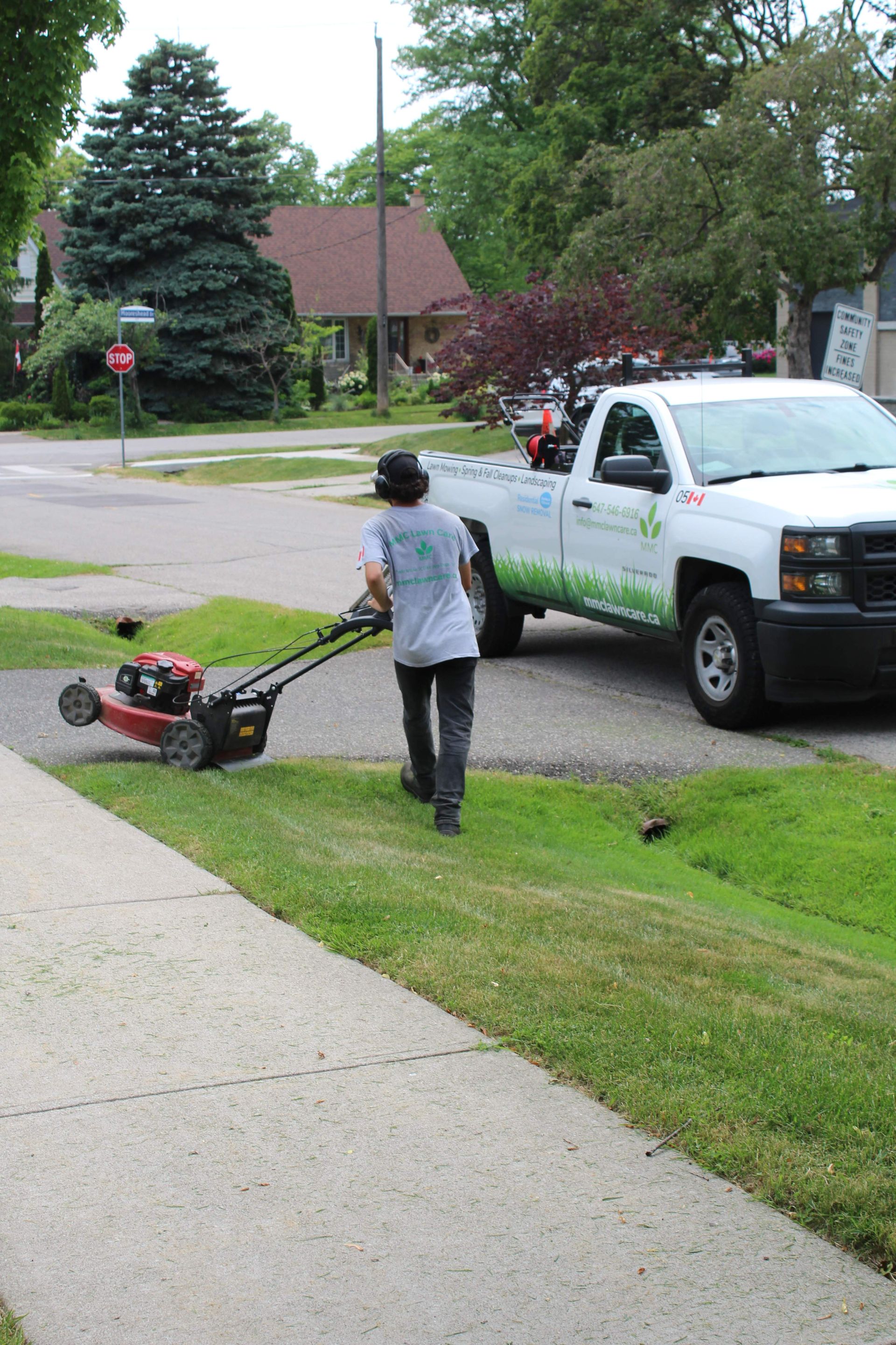 Person using a lawn mower near a white pickup truck on a suburban street sidewalk.