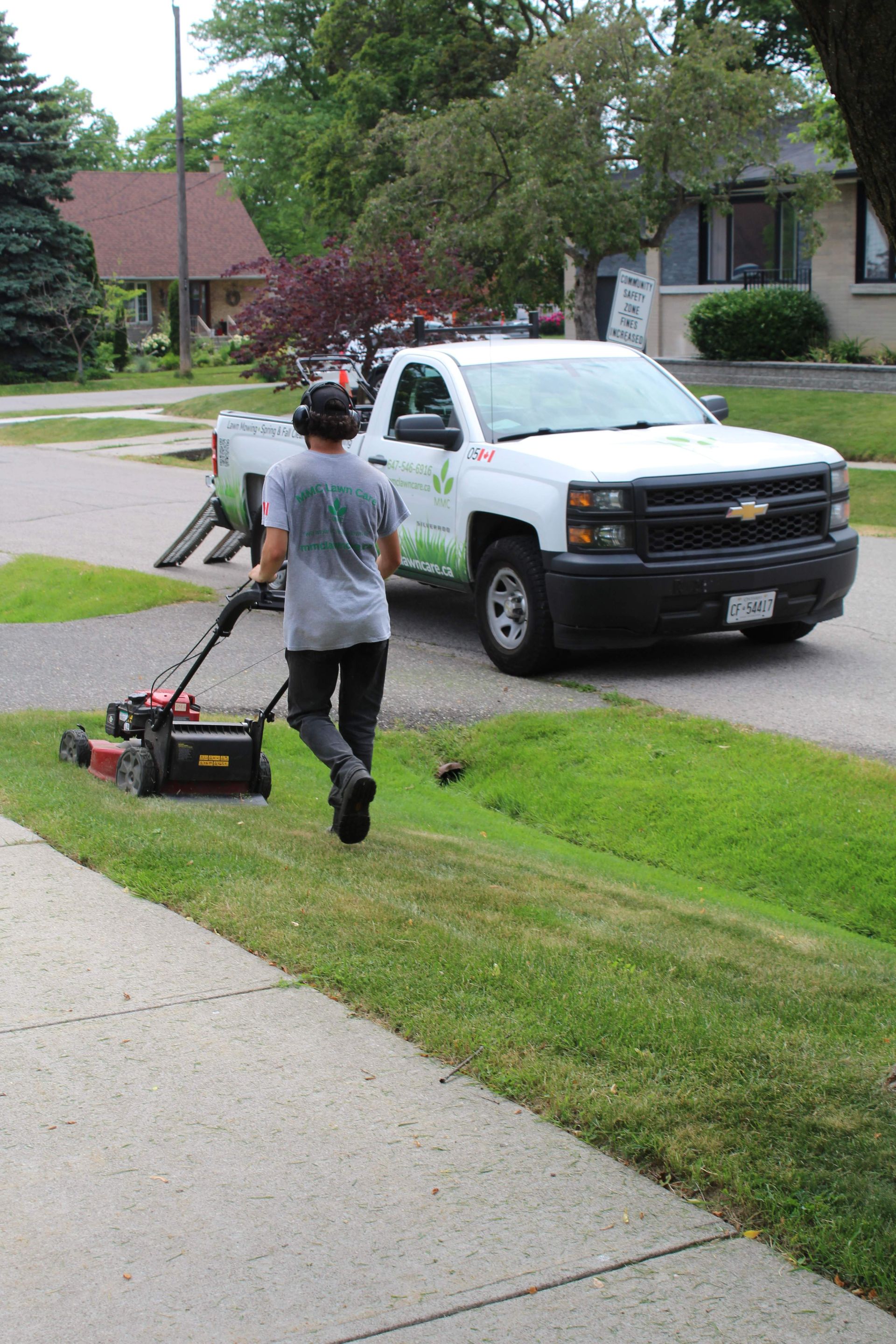 Two workers using lawn mowers on a grassy roadside near a white pickup truck in a neighborhood.