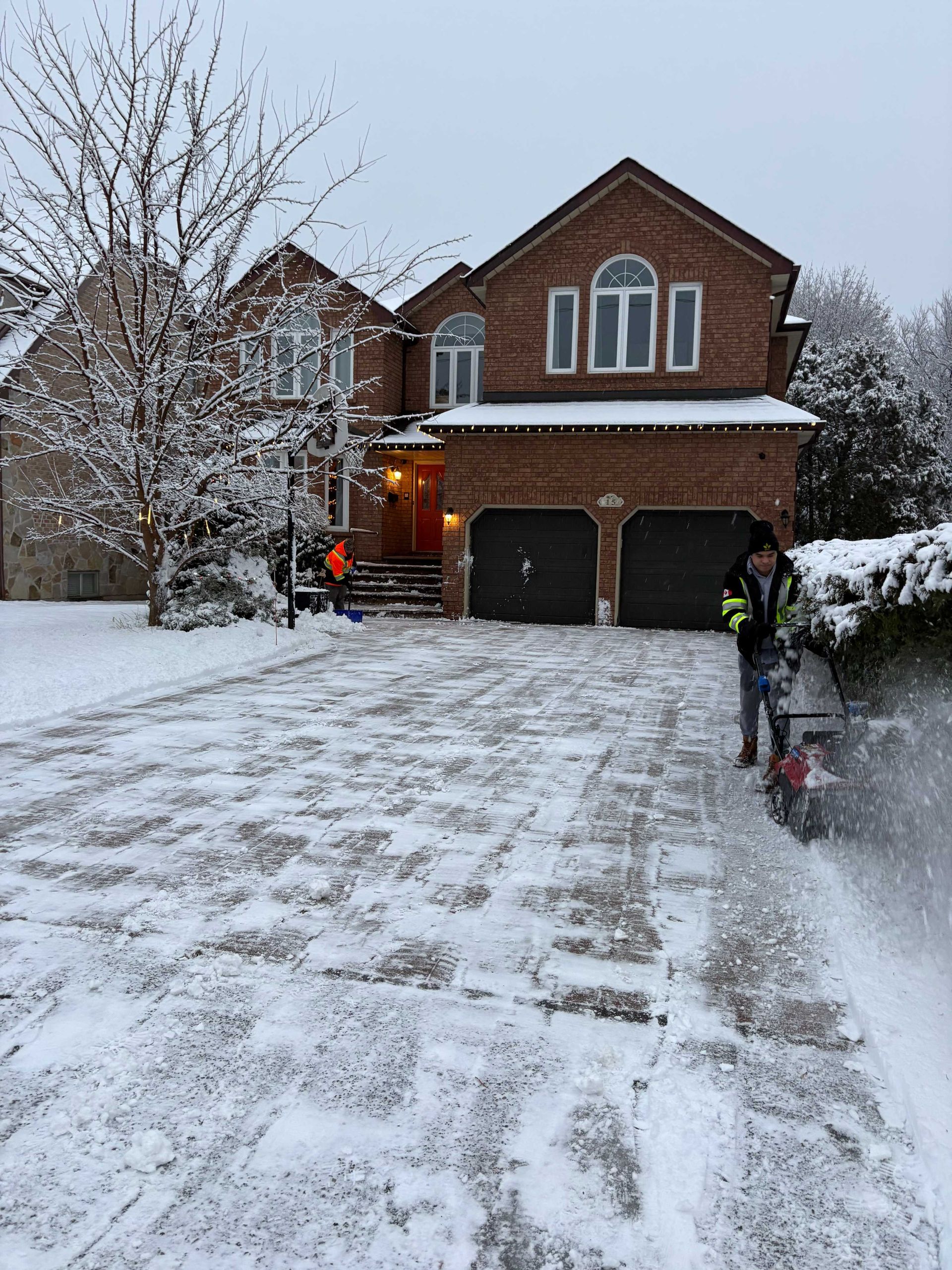 Two people clearing snow from a driveway in front of a brick house. One uses a snowblower.