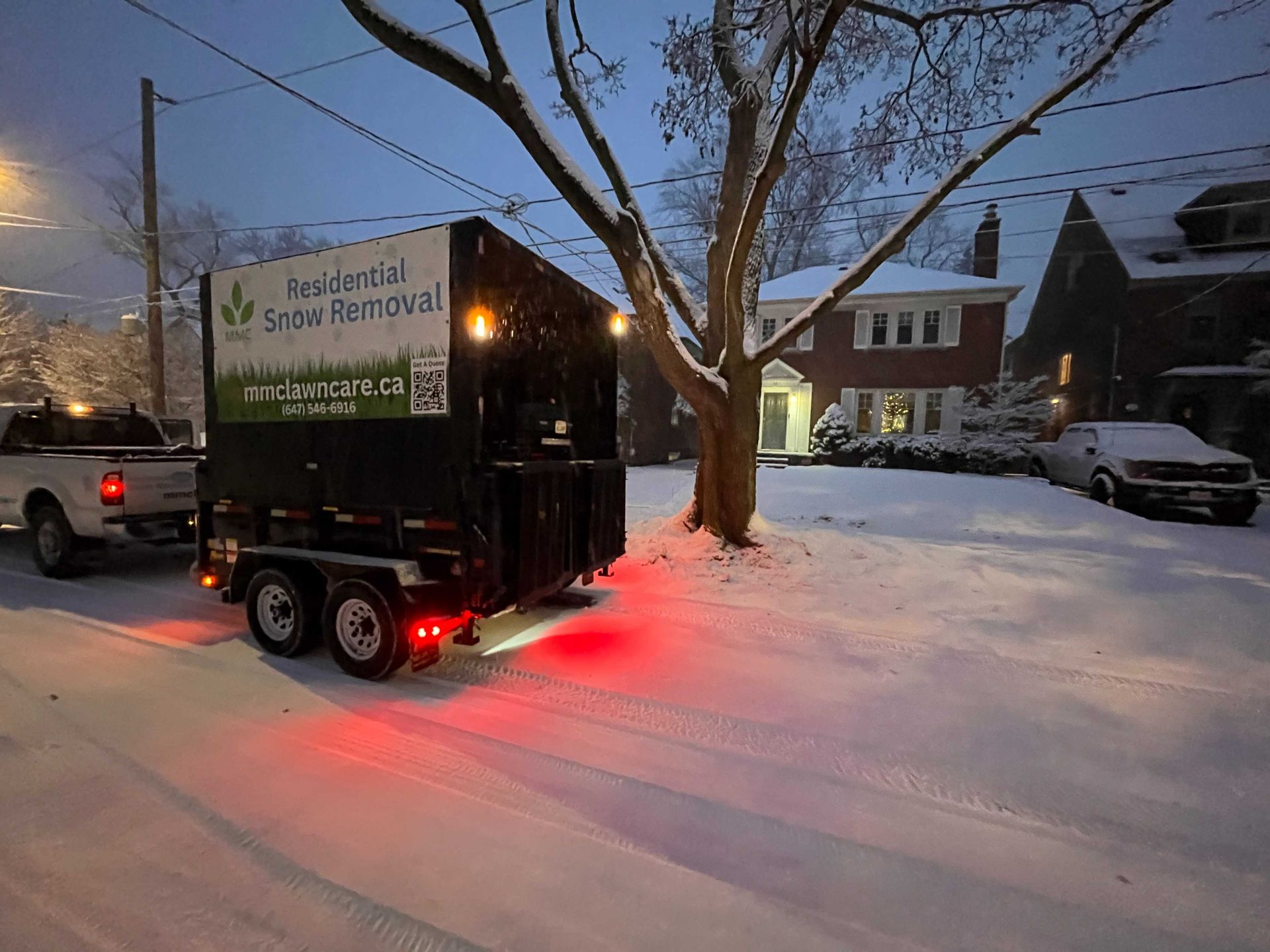 Snow removal trailer hitched to a truck on a snow-covered residential street at dusk.