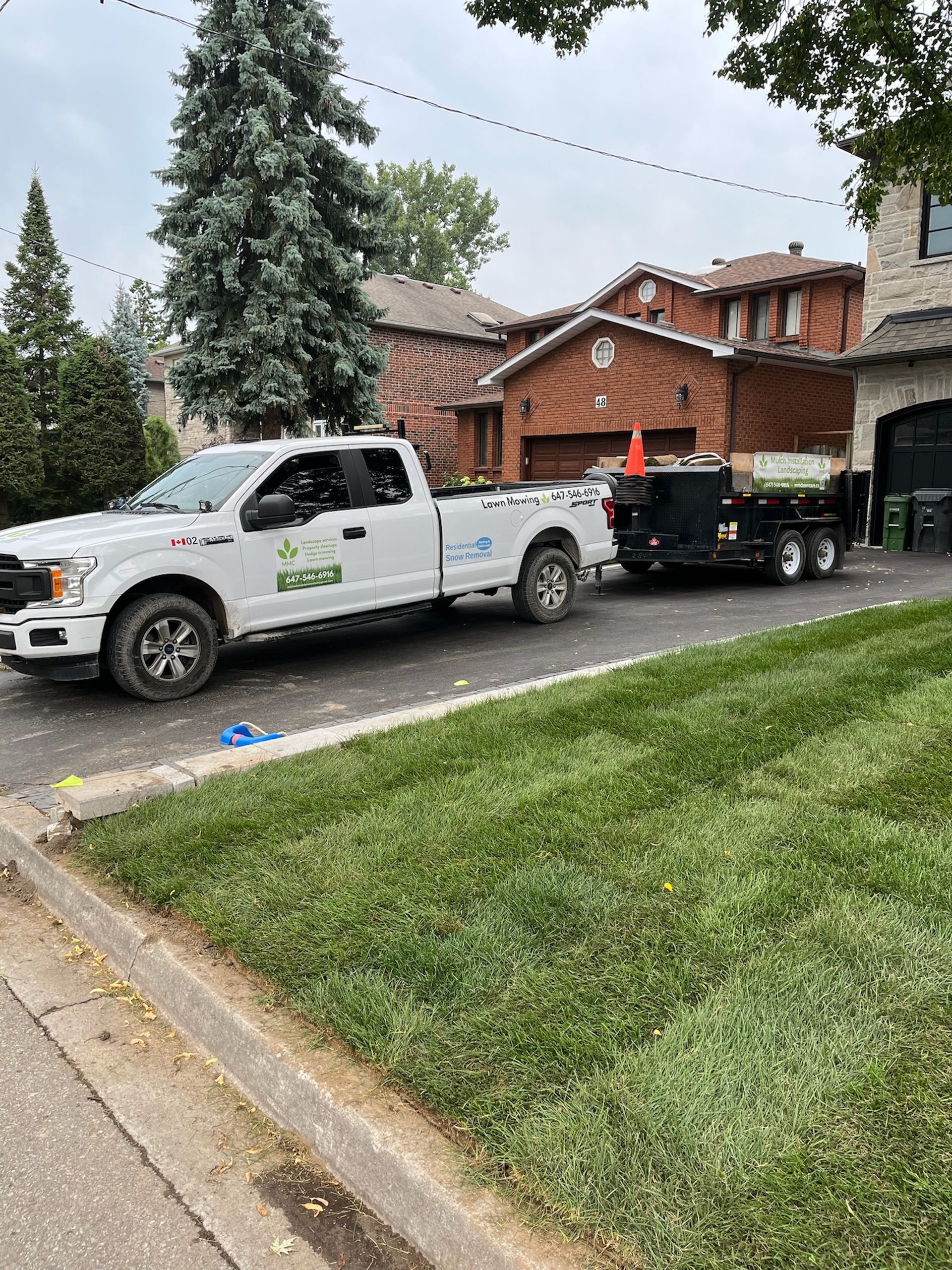 White pickup truck towing a trailer with landscaping equipment on a residential street. Green grass and houses in background.