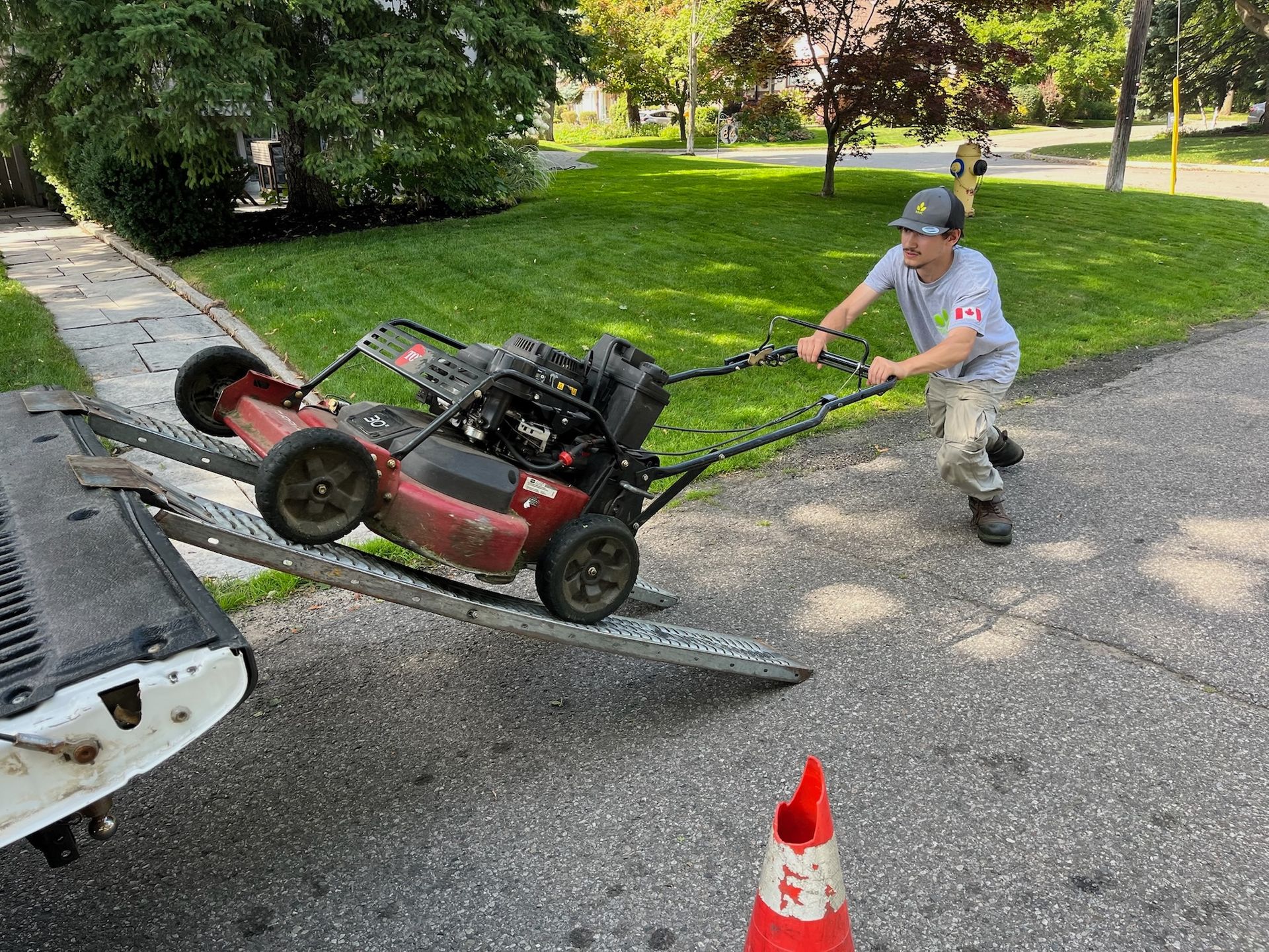 Man pushing a red lawnmower down a ramp into a truck bed on a driveway.