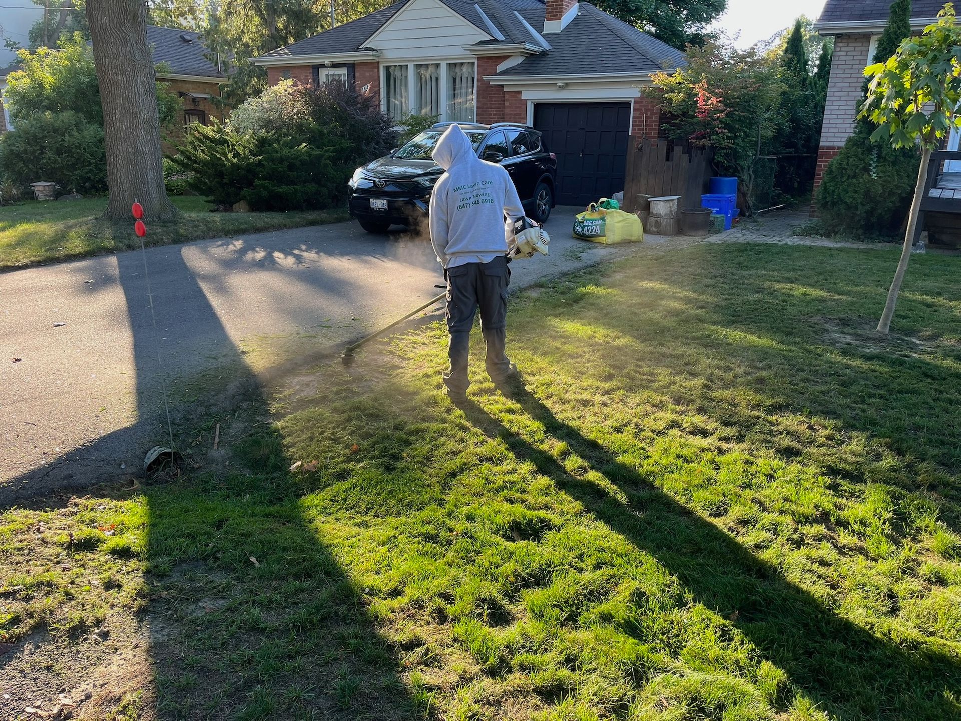 Person in a hooded sweatshirt sprays a lawn with a long shadow. A house with a driveway and car is in the background.