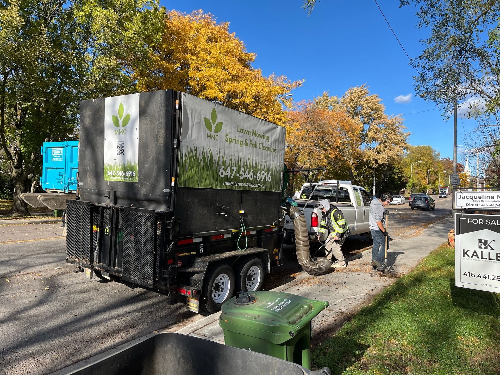 A truck with a leaf vacuum trailer, collecting leaves on a street lined with trees in autumn.