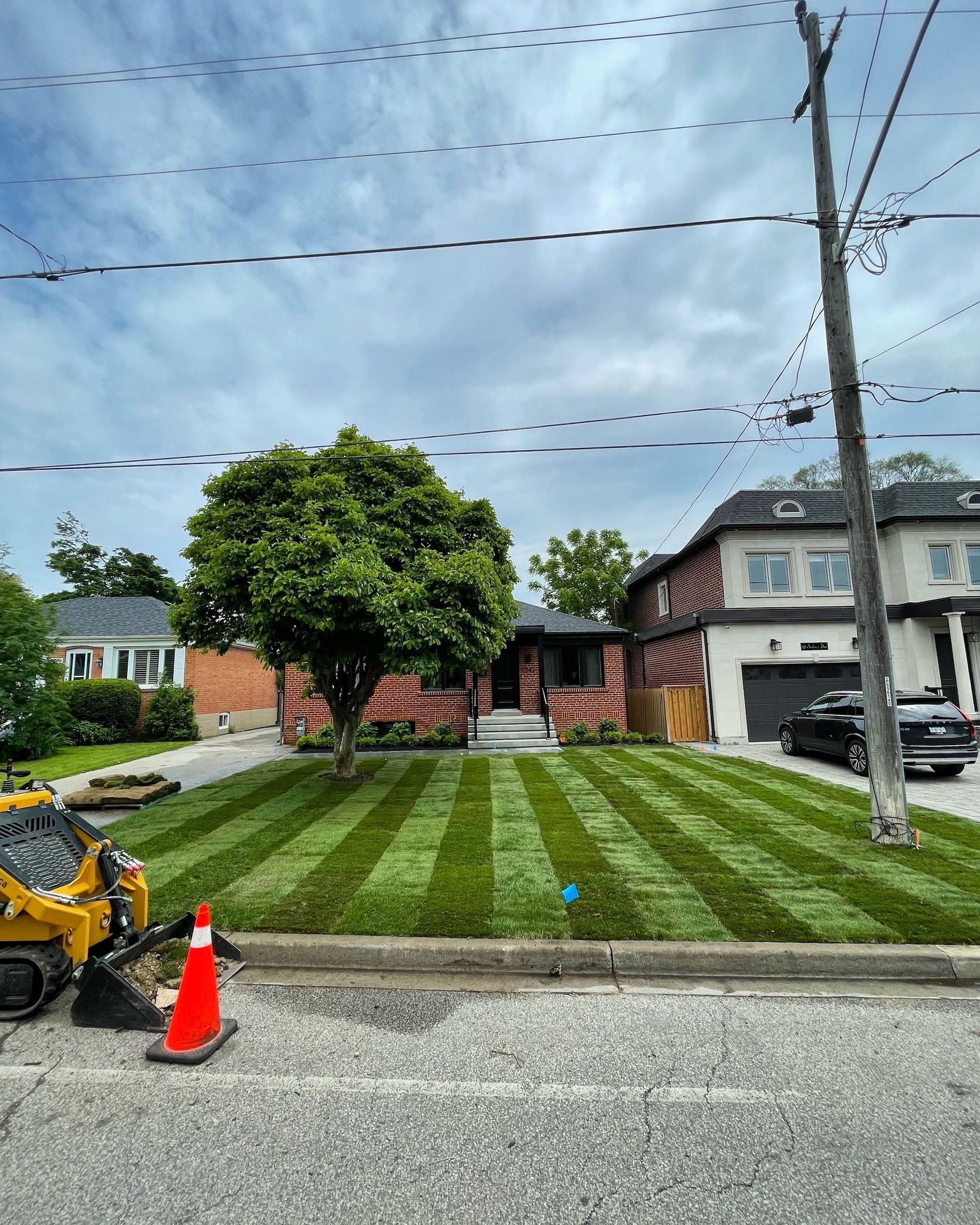 Green lawn with striped mowing pattern in front of a brick house. Construction equipment, power lines, and cloudy sky present.