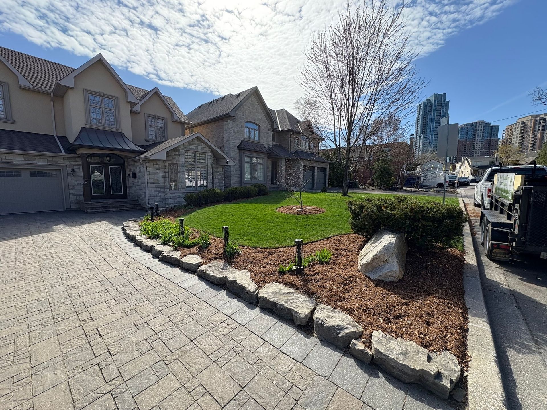 Stone house with brick driveway, small lawn, mulch, and rock border, on a city street under a blue sky.