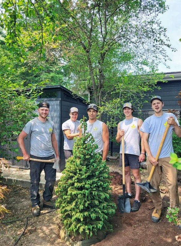 Five people with shovels and tools stand in a garden near a tree and a shed.
