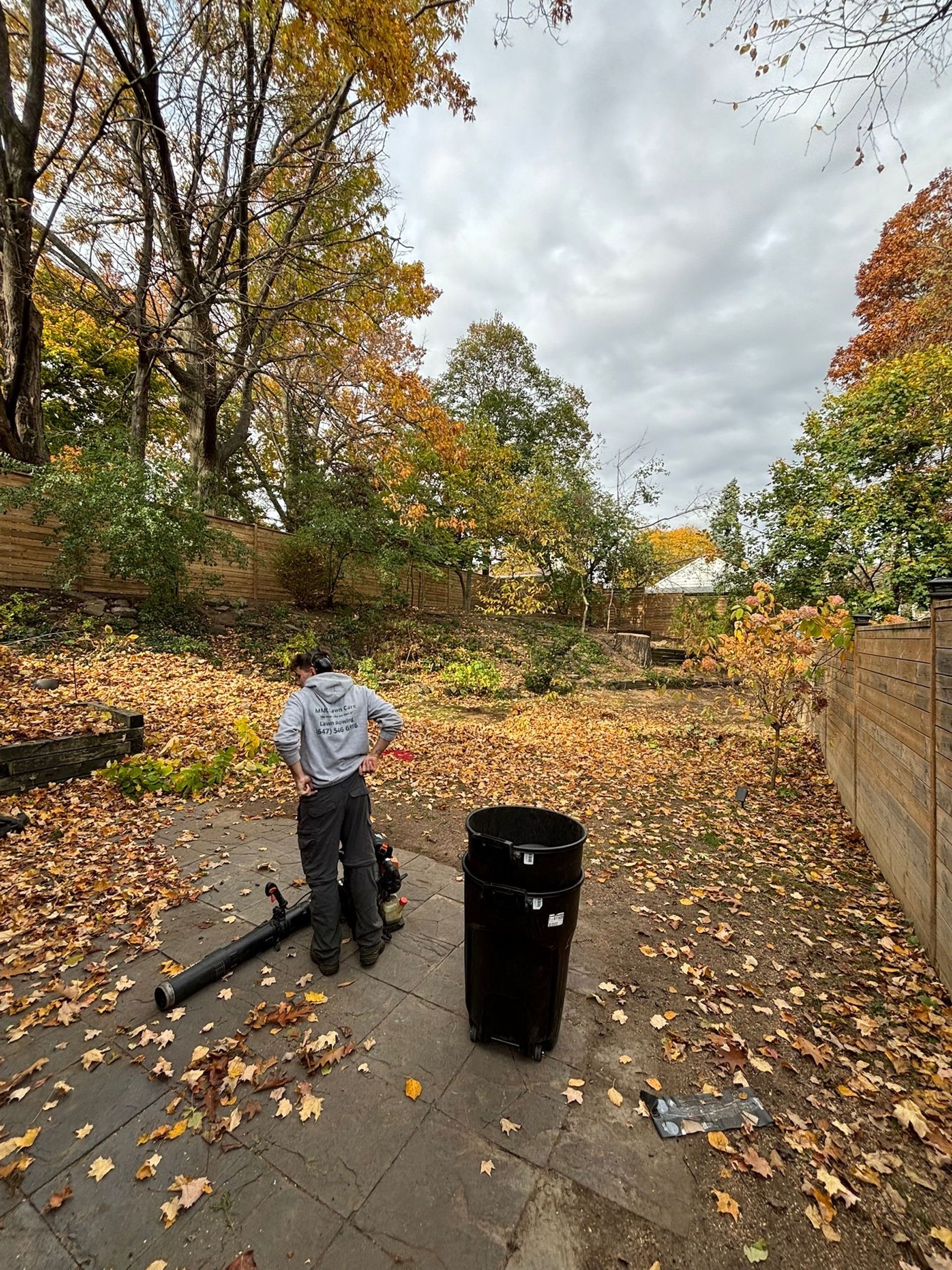 Person working in an autumn backyard, raking leaves near a black trash bin and a fence.