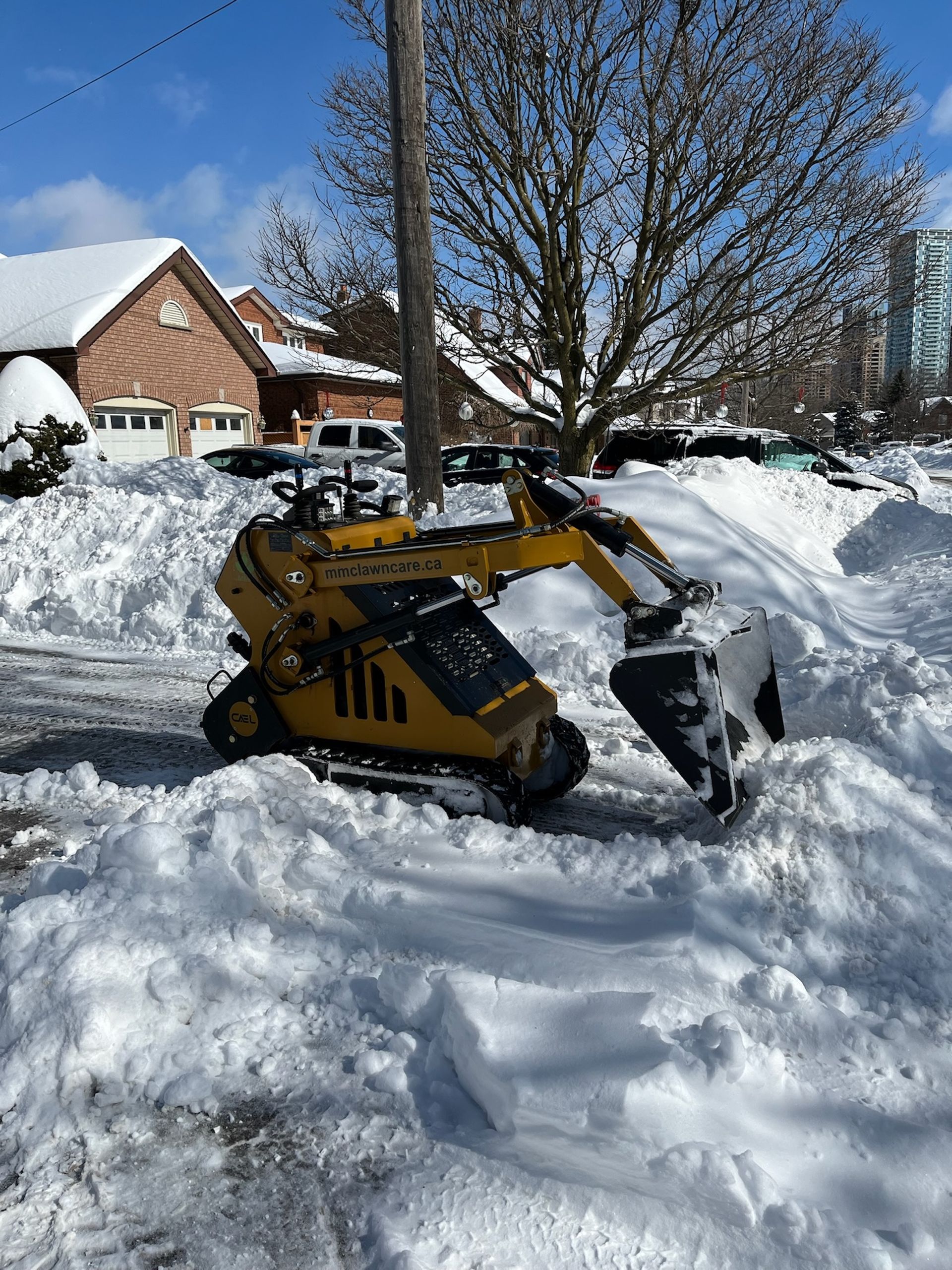 Yellow mini-excavator clearing snow from a street in front of houses on a sunny day.