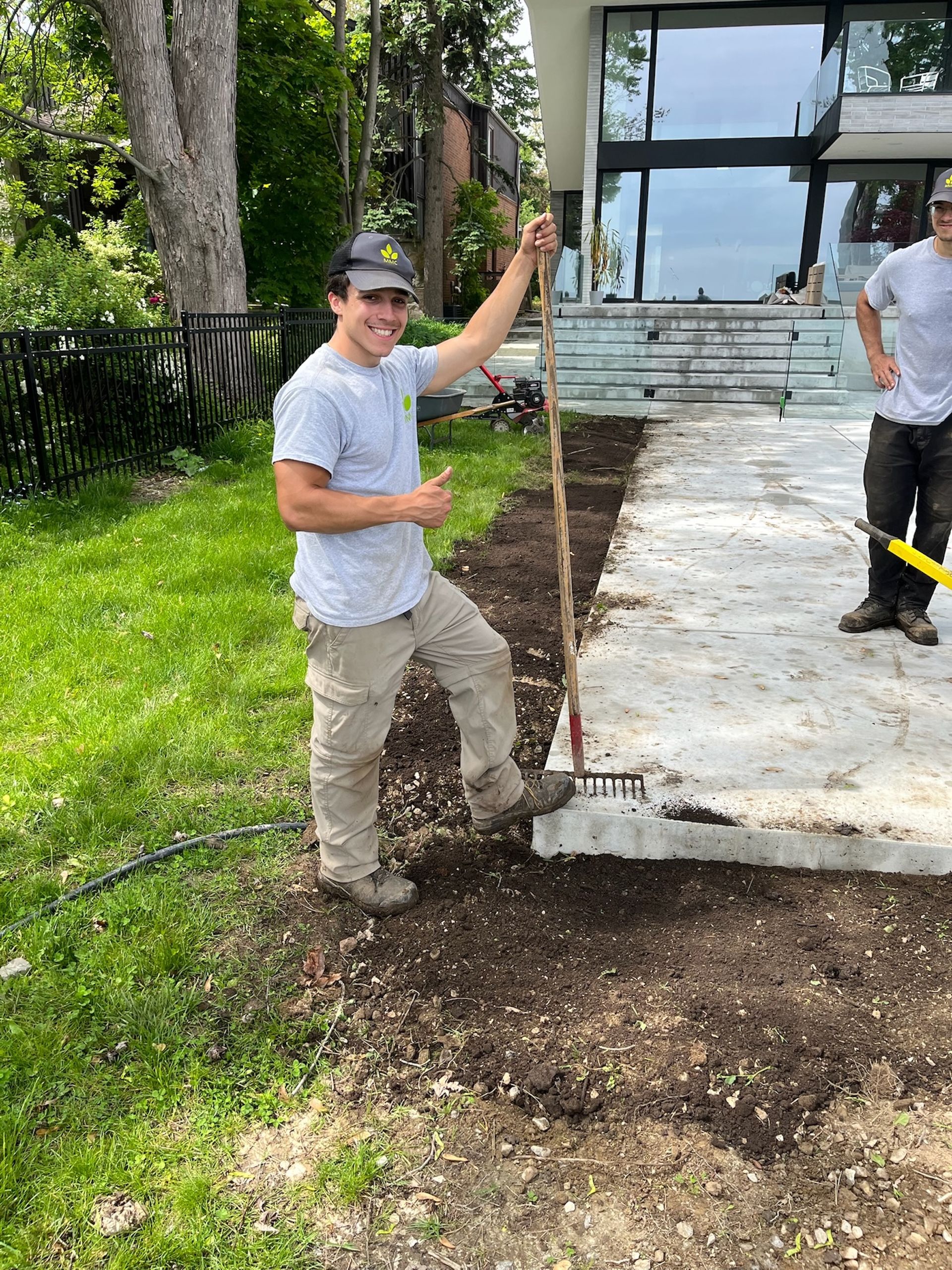 A construction worker smiles, holding a rake over a concrete patio under construction.