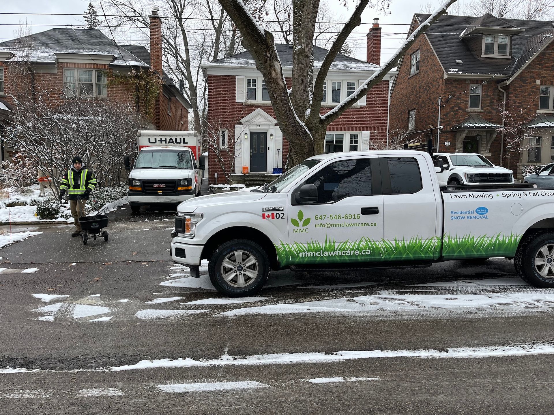A white pickup truck with logo parked on a snow-covered street, a worker spreading salt.