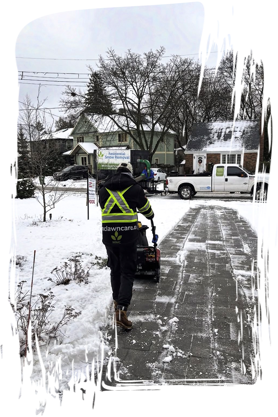 Worker clearing snow from a sidewalk beside a snow-covered street with parked trucks and houses.
