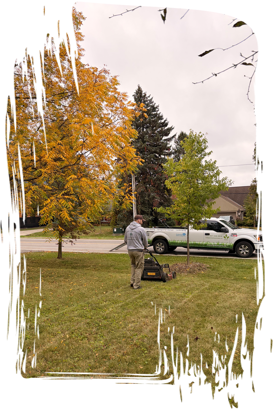 Person mowing a lawn in a suburban yard beside a white pickup truck and autumn trees.
