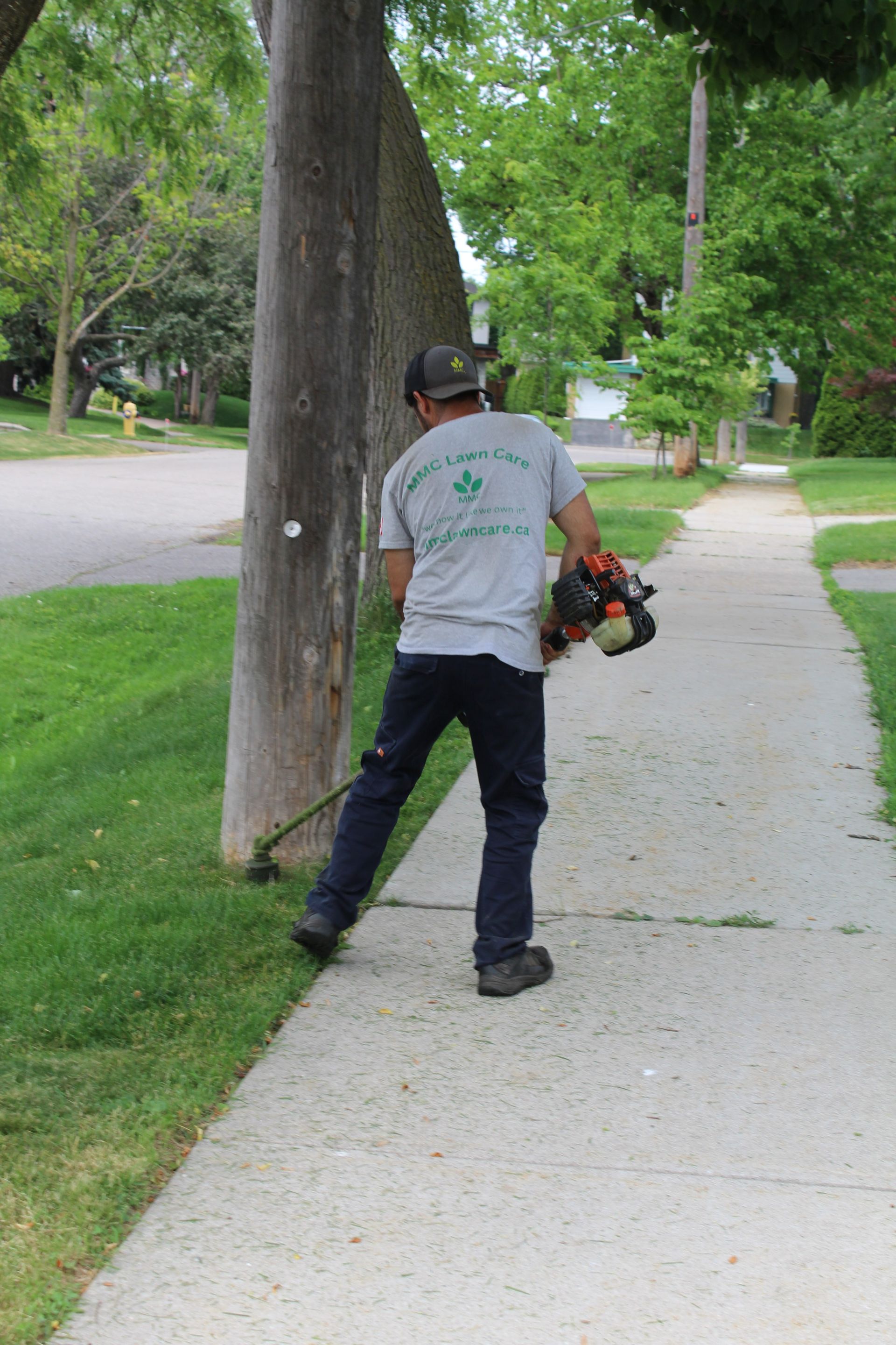 Worker trimming grass beside a sidewalk with a string trimmer in a leafy park