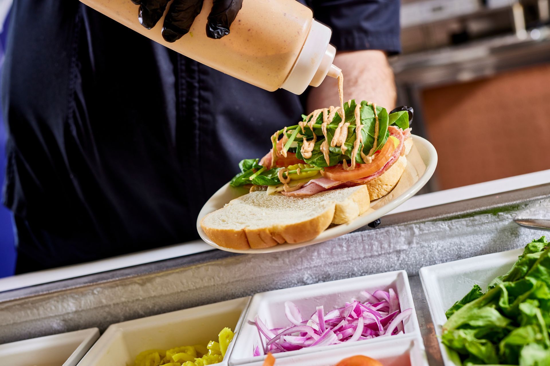 Sandwich being assembled with sauce, greens, and onions at a deli counter