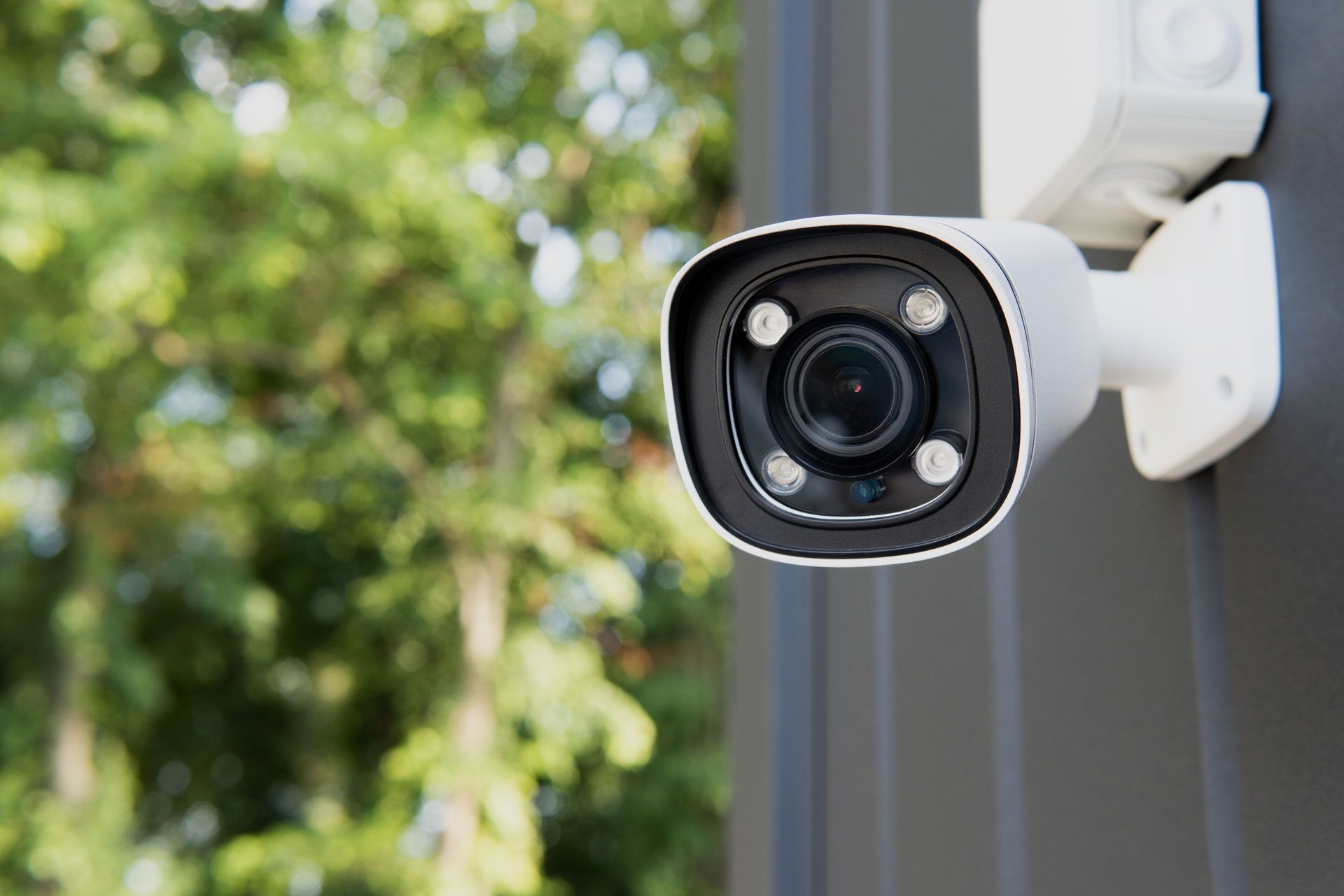 White security camera mounted on a dark surface, with a blurry green tree background.