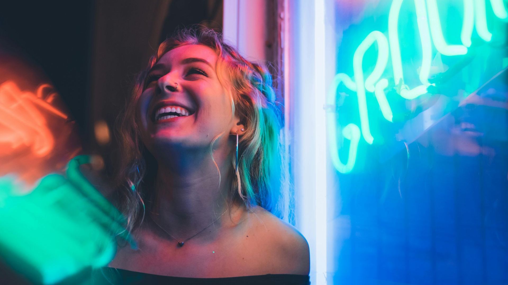 A woman is laughing in front of a neon sign.