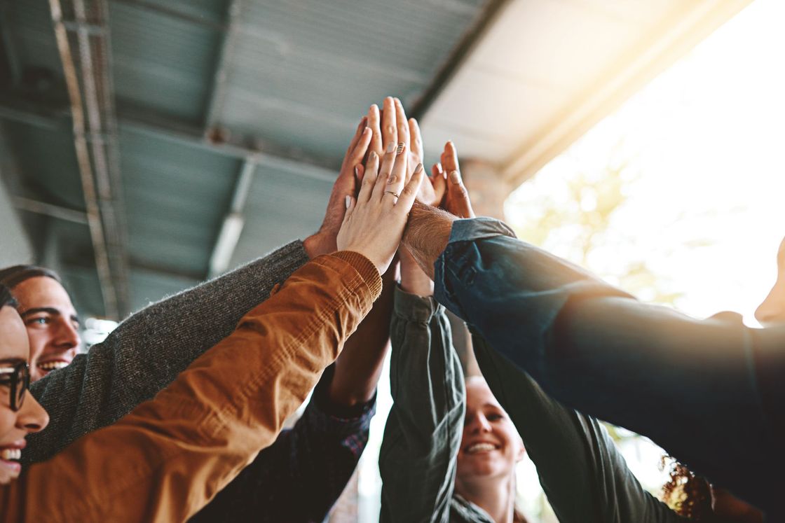People stacking hands together in a team huddle indoors, with sunlight streaming in behind them