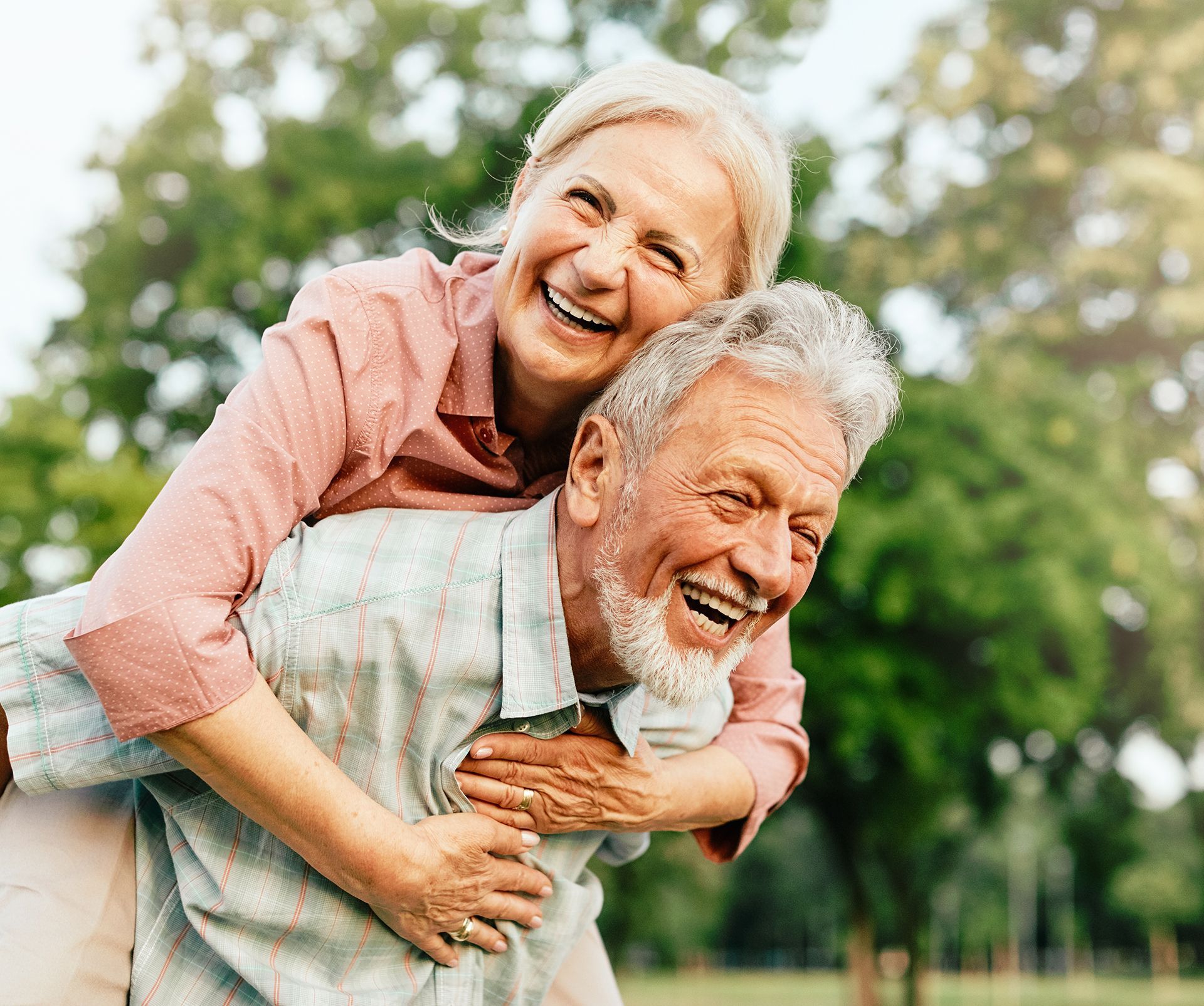 Smiling older adult couple; woman piggybacking on man outdoors in a park.