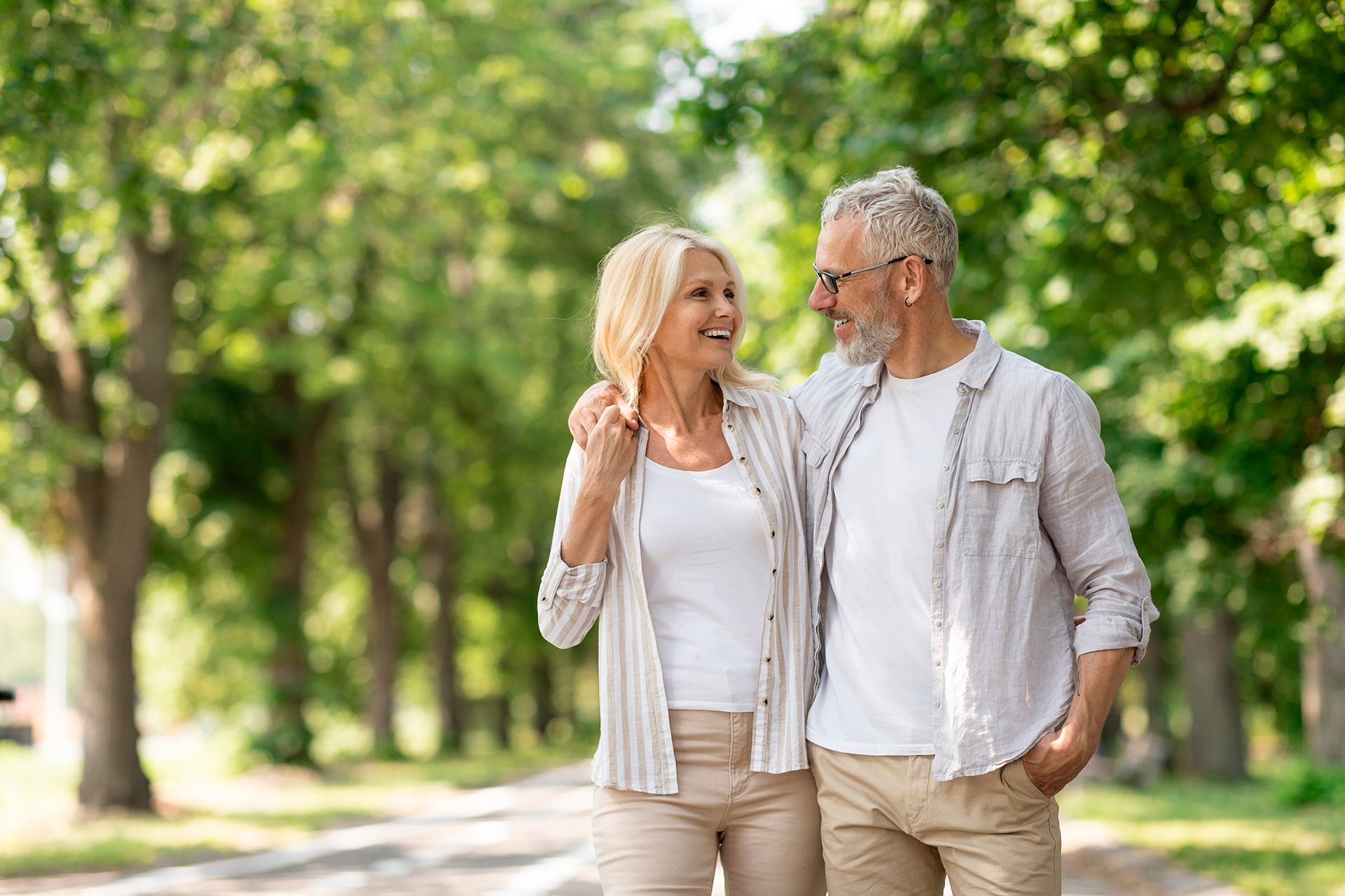 An older couple strolls arm in arm in a park, smiling at each other.