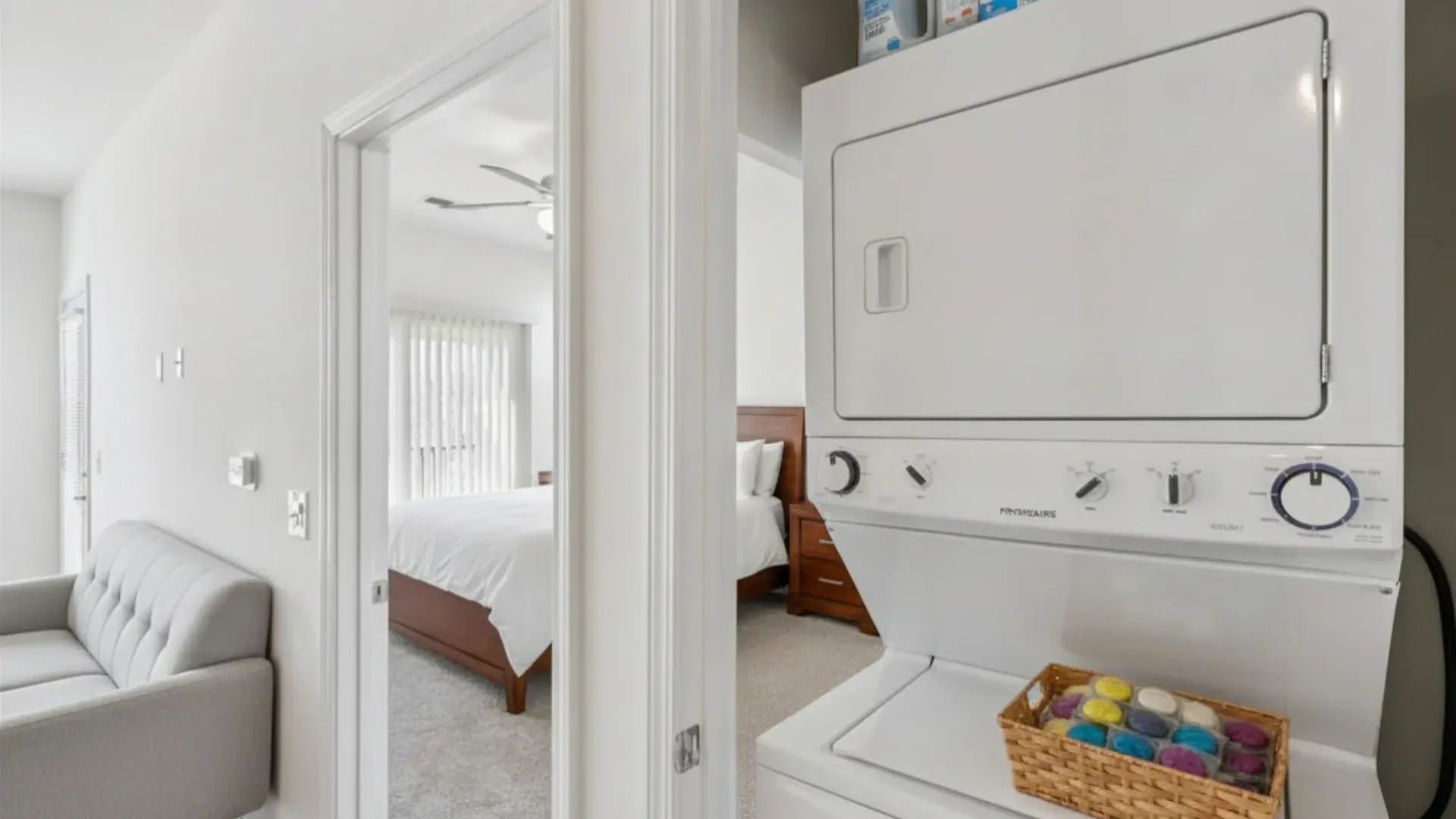 Stacked washer and dryer with a basket of laundry pods, open doorway to bedroom with bed and ceiling fan at Catherine 36 Apartments for rent in Charlotte, NC