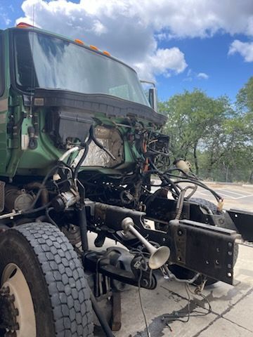 Damaged green truck front end with exposed engine and detached bumper on a roadside under a blue sky