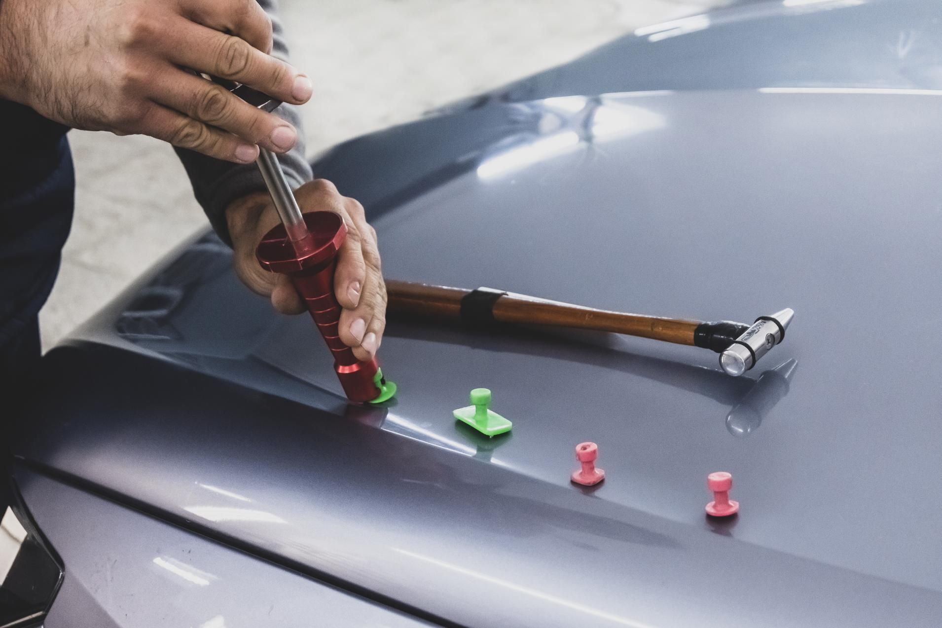Hands repairing a gray car hood with a suction cup tool and small colored dent tabs